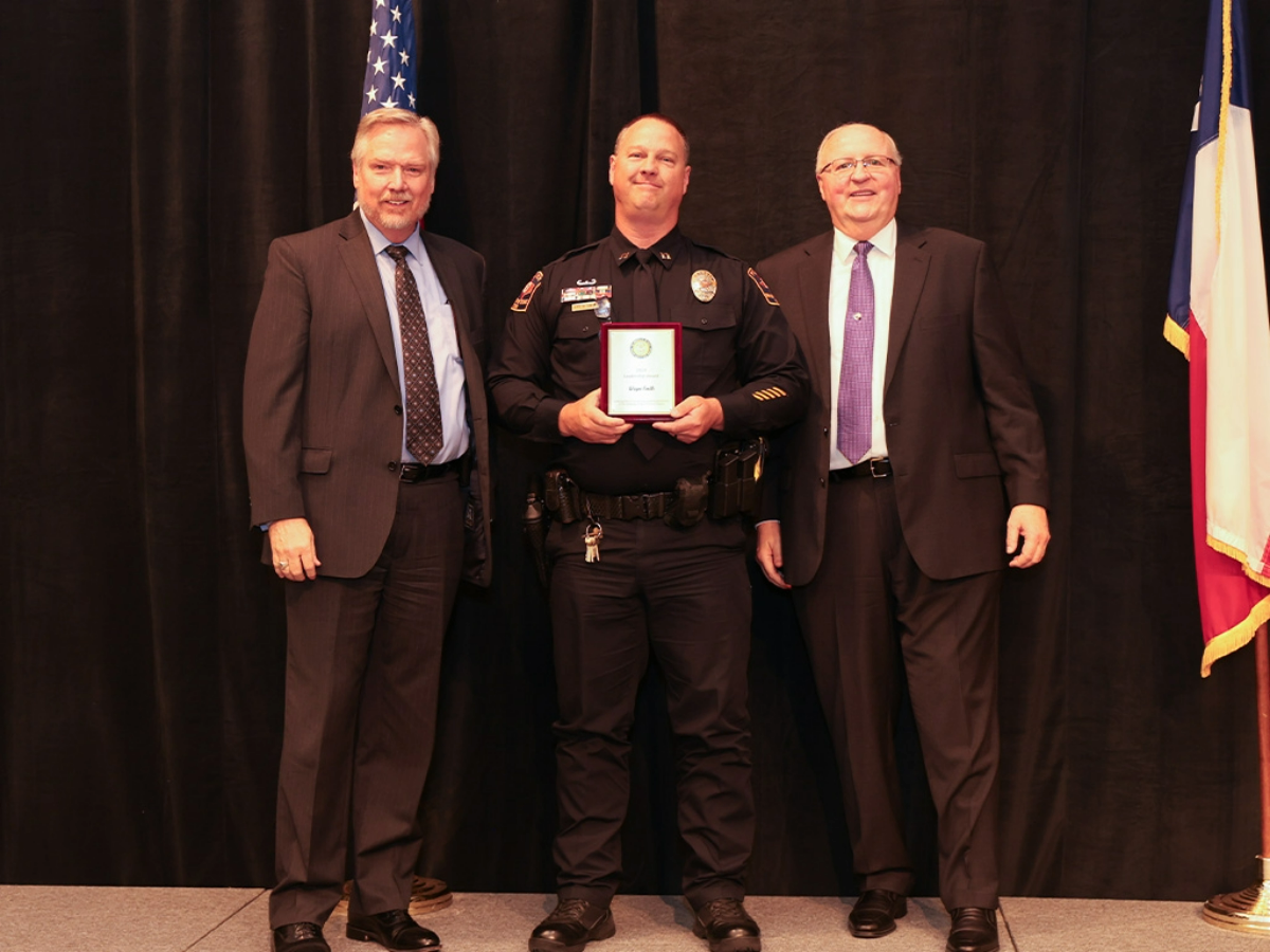 UT Police awards ceremony—uniformed officer stands between two suited administrators, holding a plaque on a stage with a black backdrop; U.S. and Texas flags frame the group.